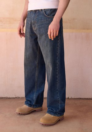 Persona con vaqueros azul oscuro holgados y botas de trabajo marrón claro de pie sobre un suelo de hormigón frente a una pared beige.