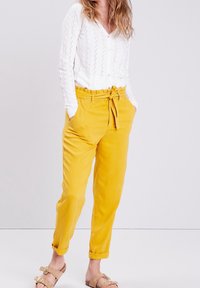 Žluté kalhoty s vysokým pasem, se zavazovacím pásem, zúženými nohavicemi a bočními kapsami. Kombinované s bílou blůzou s dlouhým rukávem a texturou.
