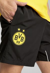 Sorte sportsshorts med et gult emblem med "BVB 09" og to stjerner. Materialet er glat, og shortsene har sidelommer med hvide accenter.