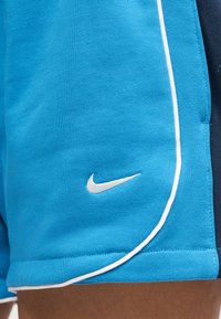 Μπλε αθλητικά σορτς με λευκή λεπτομέρεια και μικρό λευκό λογότυπο Nike Swoosh κοντά στο τελείωμα στον μηρό ενός ατόμου.