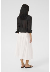 Haut noir semi-transparent à manches longues, jupe midi blanche à taille élastique, sandales plates noires ; texture lisse et design fluide avec une esthétique minimaliste.