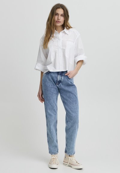 Camisa blanca corta con botones y un bolsillo, combinada con jeans de tiro alto en azul desgastado y zapatillas de lona beige. Diseño simple, ajuste relajado.