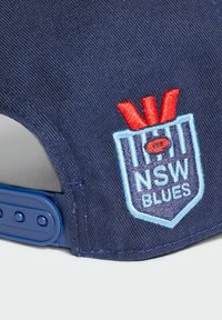 Plava pamučna kapa s izvezenim logotipom NSW Blues uz crvene akcenti, dizajn u obliku štita i trakom za podešavanje s pritiskom.