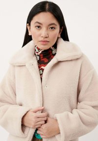 Flauschiger pinker Mantel mit breitem Kragen, Druckknöpfen und locker sitzenden Ärmeln. Über einem mehrfarbig gemusterten Rollkragenpullover getragen.