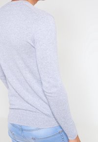 Jasnoszary sweter z fakturowanym wzorem dzianiny, długimi rękawami i lekko zaokrąglonym dołem, zestawiony z niebieskimi dżinsami.