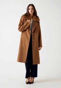 Brun uldfrakke med falsk pelskrave, enkeltradet design og to forlommer, kombineret med stribet top og bukser med vide ben.