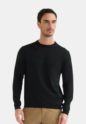 Uomo con capelli corti e scuri, che indossa un maglione nero girocollo e pantaloni beige, in piedi davanti a uno sfondo chiaro e semplice.