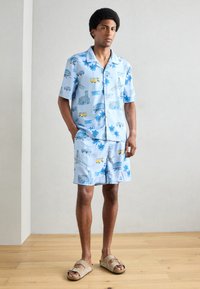 Ensemble composé d'une chemise à manches courtes bleu clair et d'un short, orné de divers motifs illustrés, y compris des palmiers et des véhicules. Porté avec des sandales beiges.