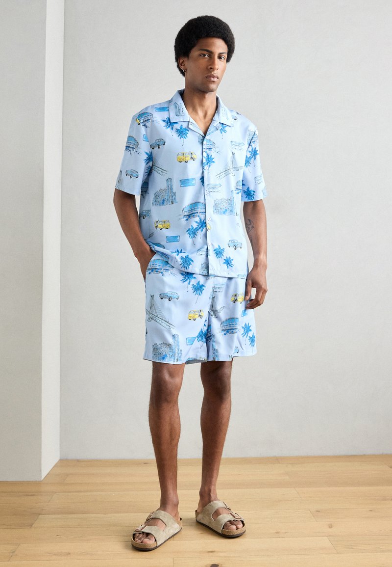 Ensemble composé d'une chemise à manches courtes bleu clair et d'un short, orné de divers motifs illustrés, y compris des palmiers et des véhicules. Porté avec des sandales beiges.
