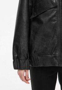 Veste en cuir noir à coupe ample, avec détails de poignets élastiques et surface texturée. Fermeture éclair et poches latérales visibles.