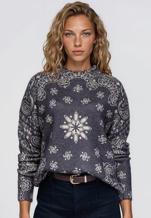 Une femme aux cheveux ondulés porte un pull gris anthracite aux motifs cachemire et floraux couleur crème, un jean foncé et une ceinture marron sur un fond clair.