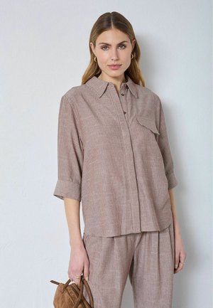 Žena v svetlohnedom pásikovanom tričku a zodpovedajúcich nohaviciach drží malú hnedú semišovú kabelku na bielom pozadí.