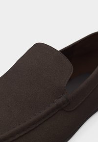 Mocassin en daim marron foncé avec couture visible le long du bord supérieur, mettant en valeur une texture lisse et un design sans lacets.