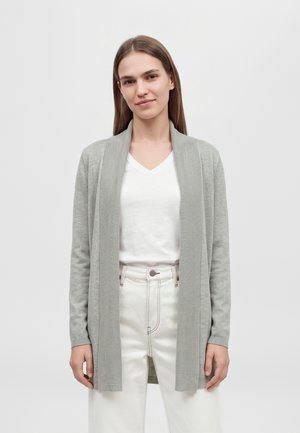 Une femme porte un long cardigan gris clair sur un haut blanc à col en V et un pantalon blanc, devant un fond de studio uni.