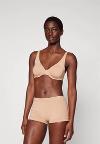 Žena s krátkými vlasy v béžové podprsenkové podprsence s kosticemi a odpovídajícím vysokým spodním prádlem, stojící na světlém jednobarevném pozadí.