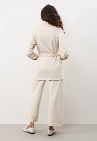 Beige långärmad jacka med bälte i midjan och klassisk krage, i kombination med vida byxor, som har en slät textur och minimalistisk design.