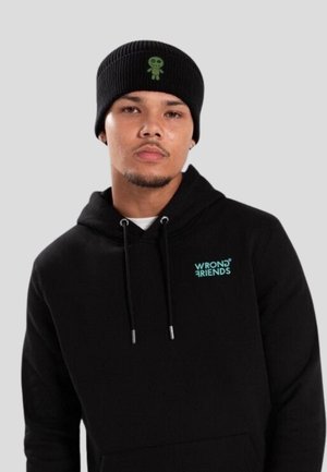 Mann trägt eine schwarze Strickmütze mit grünem Alien-Patch und einen schwarzen Kapuzenpullover mit türkisfarbenem "WRONG FRIENDS" Logo auf der Brust, hellgrauer Hintergrund.
