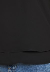 Black blouse with a smooth texture, featuring a rounded hem and stitching details. Paired with light blue denim jeans.