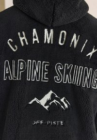 Juodas vilnonis džemperis su siuvinėtu tekstu "Chamonix Alpine Skiing", su kalno dizainu ir detalėmis "Off-Piste" apačioje.