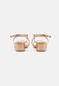 Metallic roséguld klacksandaler med två ankle straps och en strukturerad yta, med en rund blockklack och öppen tådesign.
