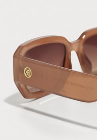 Gafas de sol de plástico marrón con un marco grueso, lentes oscuros y un logo dorado en el lateral. Textura suave, diseño moderno.