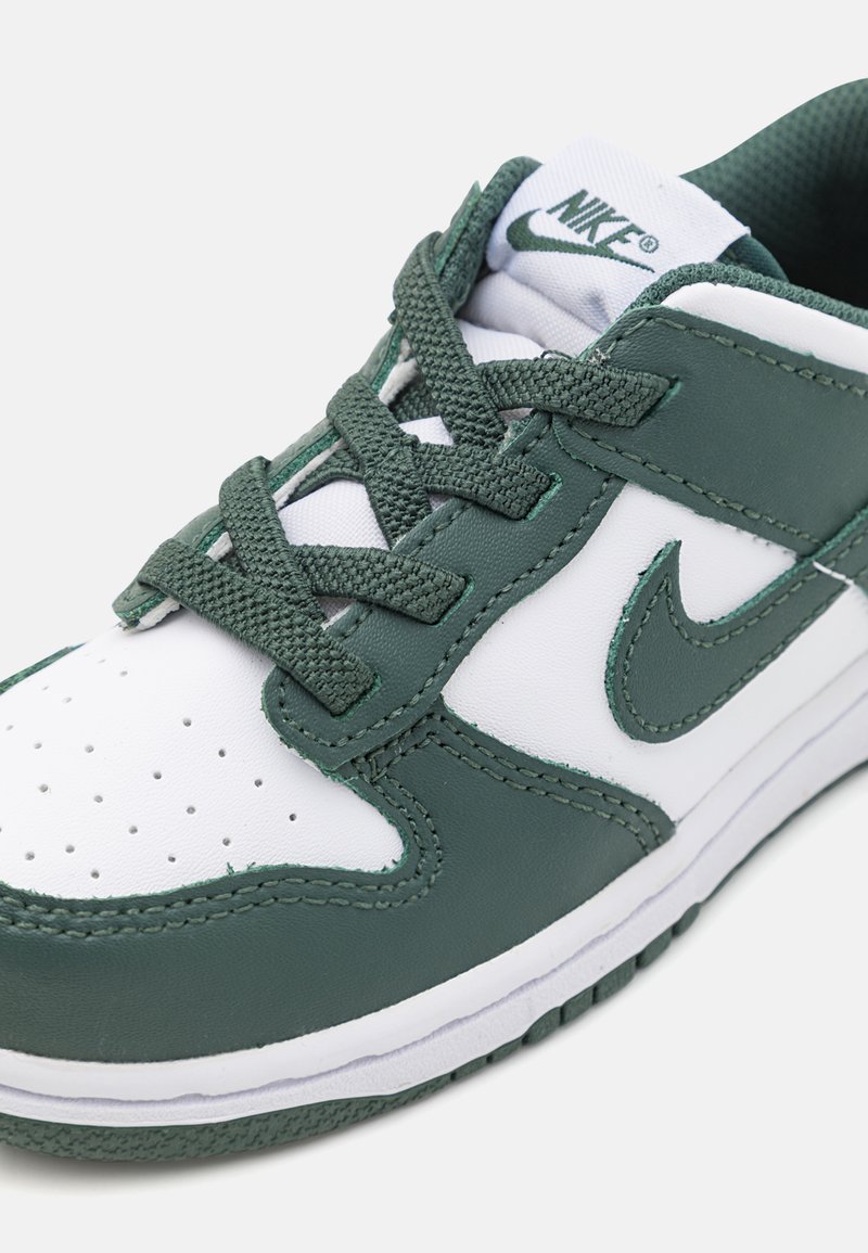 Zapatillas Nike con parte superior de cuero blanco y acentos sintéticos verdes, puntera perforada y cordones verdes texturizados sobre una suela blanca.