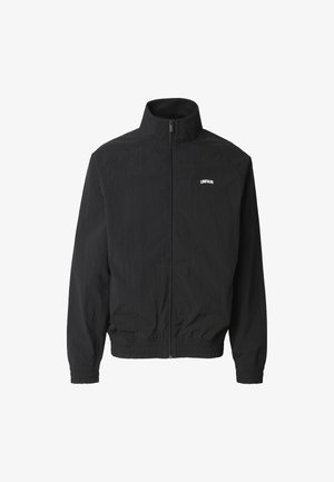 Musta vetoketjullinen takki, jossa korkea kaulus, joustavat hihansuut ja vyötärönauha, vasemmassa rinnassa pieni valkoinen "UNFAIR"-teksti.