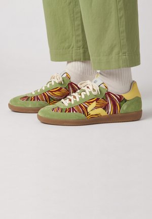 Baskets basses en daim vert, motif feuilles multicolores, semelles couleur cuir clair, lacets blancs, portées avec des chaussettes blanches et un pantalon vert court sur fond clair.