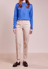 Osoba ubrana w niebieską koszulę z długim rękawem, beżowe dopasowane spodnie oraz ciemnobrązowe mokasyny stoi na tle jednolitym.