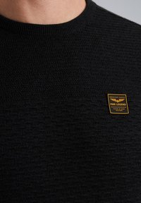 Crni pleteni dukserica s teksturiranim dizajnom i malom pravokutnom žutom oznakom branda na prsima s bijelim tekstom i logotipom.