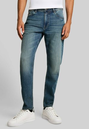 Mann, der blaue ausgewaschene Jeans und weiße Turnschuhe trägt, steht vor einem einfarbigen hellgrauen Hintergrund.