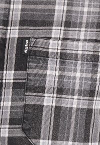 Camicia a scacchi bianca e nera con taschino sul petto, caratterizzata da un tessuto a trama e motivi a strisce diagonali. Inclusa etichetta con la scritta "tigha".
