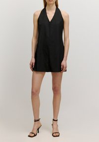 Schwarzes Neckholderkleid, knielang, aus strukturiertem Stoff. Mit tiefem V-Ausschnitt und sauberem, minimalistischen Design. Getragen mit schwarzen, strappy High Heels.