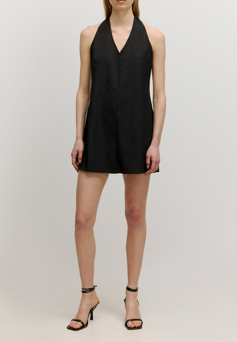 Schwarzes Neckholderkleid, knielang, aus strukturiertem Stoff. Mit tiefem V-Ausschnitt und sauberem, minimalistischen Design. Getragen mit schwarzen, strappy High Heels.