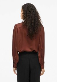 Brūna satīna blūze ar garām piedurknēm un pūkainiem plecu akcentiem, apvienota ar melnām biksēm. Gluda tekstūra, brīva piegriezuma, maiga spīduma.