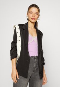 Blazer de cetim preto com design de duplo botão, combinado com uma t-shirt de alça lavanda com detalhes em renda, e jeans cinzas de cintura alta. Bolsa de ombro branca.
