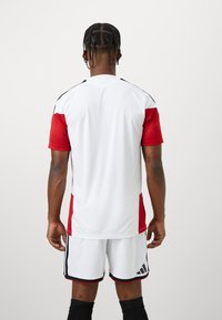 Φανέλα αθλητισμού Adidas σε λευκό και κόκκινο χρώμα, με κοντά μανίκια, κάθετες πλευρικές ρίγες και στρογγυλή λαιμόκοψη. Συνδυάζεται με λευκές βερμούδες.