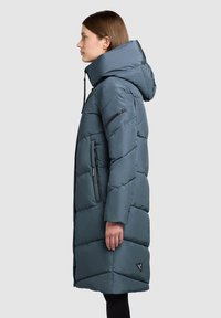Gepolsterter langer Mantel in Türkis mit hohem Kragen und Kapuze. Verfügt über ein diagonales Steppmuster, seitliche Reißverschlusstaschen und ein Logodetail.
