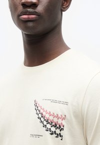 Ανοιχτό μπεζ βαμβακερό μπλουζάκι με γραφικό σχέδιο στυλιζαρισμένων αποτυπωμάτων ελαστικών σε ροζ και μαύρο, με κείμενο πάνω και κάτω.