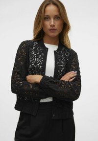 Veste bomber en dentelle noire avec des découpes florales, col rond et manches élastiquées, portée sur un top blanc. Fermeture à bouton simple.