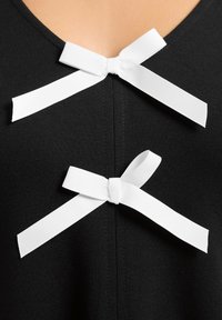 Schwarzes Kleidungsstück mit einem V-Ausschnitt, das zwei weiße Satinbänder am Halsauschnitt hat, was für Detail und Textur sorgt. Glatte Stoff-Oberfläche.