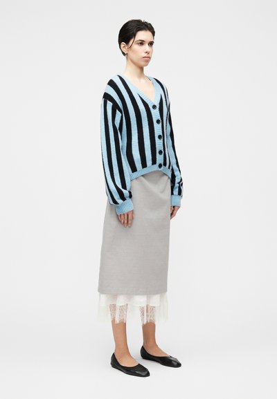 Μπλε και μαύρη ριγέ ζακέτα με V-λαιμόκοψη, κούμπωμα με κουμπιά και φαρδιά μανίκια. Συνδυασμένη με γκρι φούστα και μαύρα φλατ παπούτσια.