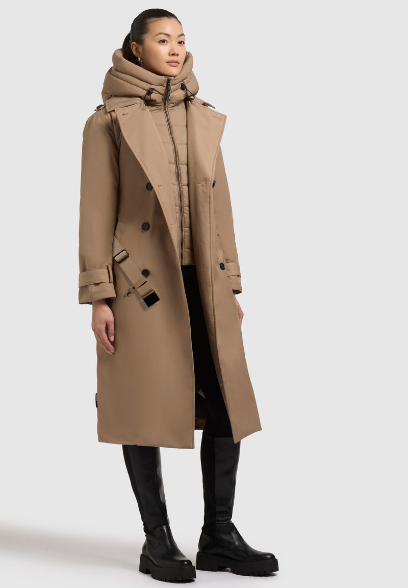 Trench coat beige con fodera imbottita e cappuccio staccabile. Presenta bottoni doppi e accentuazioni con cintura. Il modello indossa stivali neri fino al ginocchio.