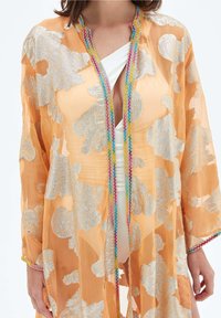 Průhledné oranžové kimono se stříbrnými květinovými vzory a multibarevnými zigzagovými detaily, nošené přes bílé plavky.