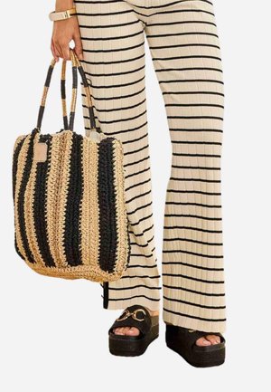 Persona que lleva pantalones crema con rayas negras, sostiene un bolso tejido a rayas negras y beige, y lleva sandalias negras de plataforma con detalles dorados.