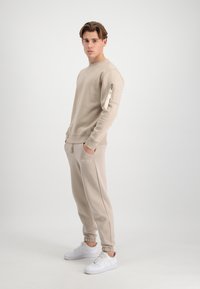 Beige sweatshirt med en lynlås lomme på venstre ærme, parret med matchende joggers. Hvide sneakers fuldender outfittet. Glat stoftekstur.