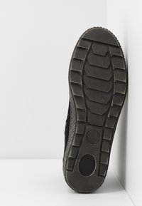 Semelle d'une chaussure noire avec des crampons profonds, posée contre un mur d'angle blanc sur une surface blanche.
