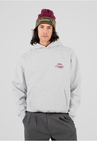 Sudadera ligera gris con un bolsillo frontal y un logotipo bordado en marrón, combinada con un gorro marrón con acentos amarillos y verdes.