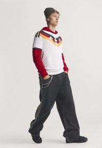 Jeune homme portant un maillot de football allemand blanc par-dessus un sweat à capuche rouge, pantalon noir oversize avec chaîne, bonnet noir et baskets noires, debout les mains dans les poches.