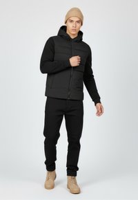 Black puffer jacket with a hood, textured upper panel, zip closure, paired with black pants and tan boots, standing on a light background.
